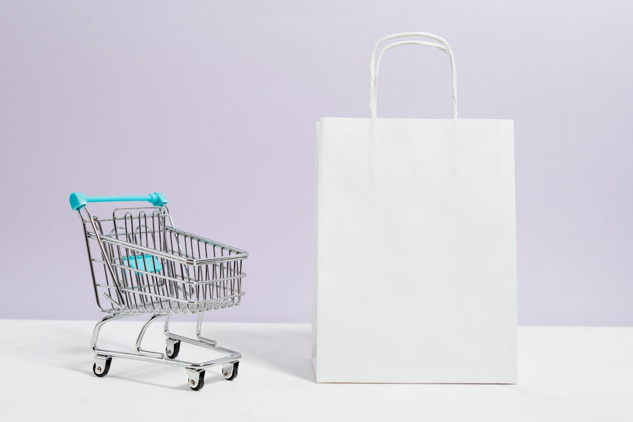 Minimalist image of a white shopping bag and mini cart on a soft pastel background.